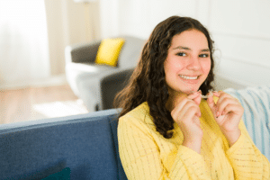 Smiling teen holding a clear orthodontic aligner while seated on a couch