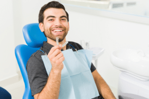 Smiling man sitting in a blue dental chair holding up a clear aligner tray