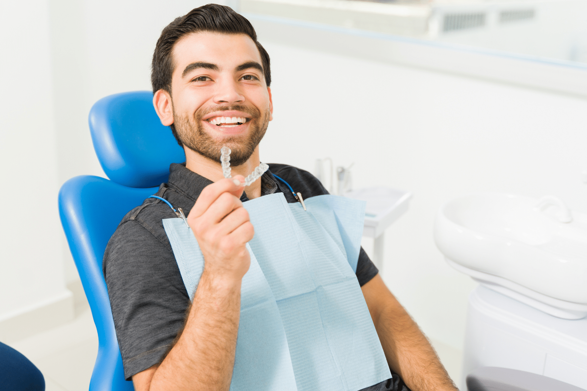 Smiling man sitting in a blue dental chair holding up a clear aligner tray